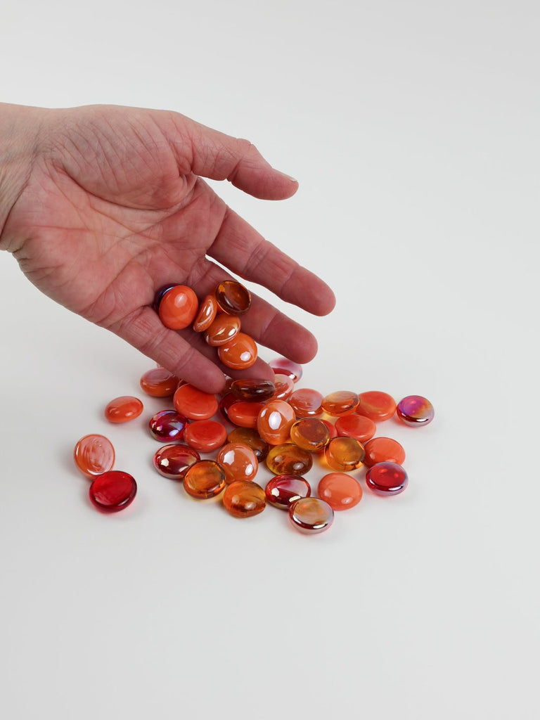 Video of orange mix glass nuggets in orange, coral and red shades with an iridescent sheen, 16–20 mm, picked up from a pile by hand and dropped back to show their size, round shape and shine