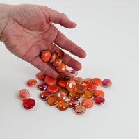 Video of orange mix glass nuggets in orange, coral and red shades with an iridescent sheen, 16–20 mm, picked up from a pile by hand and dropped back to show their size, round shape and shine